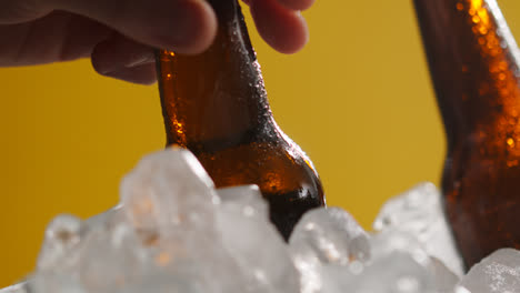 Nahaufnahme-Einer-Person,-Die-Eine-Gekühlte-Glasflasche-Mit-Kaltem-Bier-Oder-Alkoholfreien-Getränken-Aus-Einem-Mit-Eis-Gefüllten-Eimer-Vor-Gelbem-Hintergrund-Nimmt